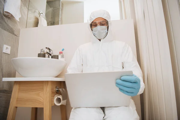 Hombre en traje de materiales peligrosos, máscara médica, guantes de látex y gafas usando el ordenador portátil en el baño cerca del lavabo - foto de stock