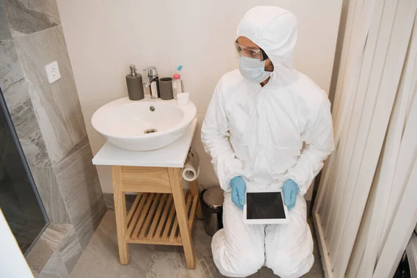 Hombre en traje de materiales peligrosos, gafas, máscara médica y guantes de látex que sostiene la tableta digital con pantalla en blanco en el baño - foto de stock