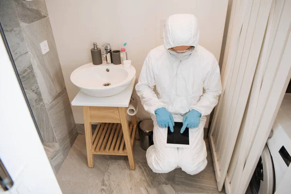 Человек в защитном костюме, медицинская маска, латексные перчатки и очки, указывающие пальцами на цифровой планшет с чистым экраном в ванной комнате — стоковое фото