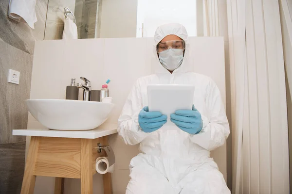 Hombre en traje de materiales peligrosos, gafas, máscara médica y guantes de látex usando tableta digital en el baño - foto de stock