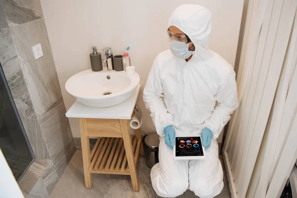 Hombre en traje de materiales peligrosos, máscara médica, guantes de látex y gafas usando tableta digital con gráficos y gráficos en el baño - foto de stock