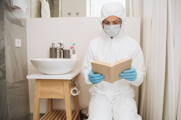 Hombre en traje de materiales peligrosos, máscara médica, guantes de látex y gafas libro de lectura en el baño - foto de stock