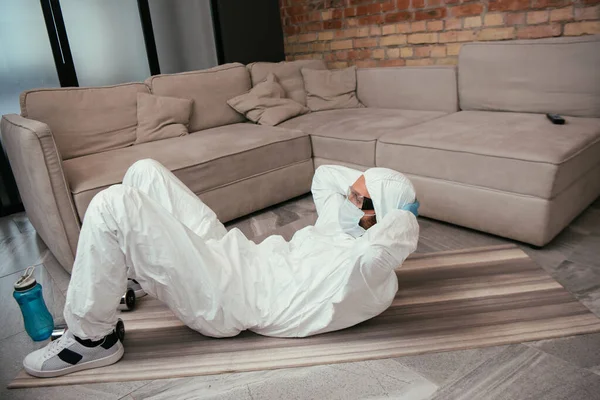 Man in hazmat suit, medical mask and goggles exercising near sports bottle in living room — Stock Photo