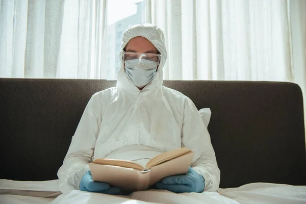 Homme en combinaison Hazmat, masque médical, gants en latex et lunettes livre de lecture au lit — Photo de stock