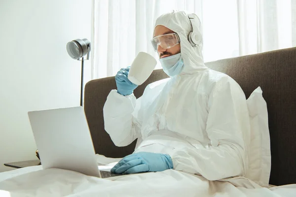 Freelancer in hazmat suit, headphones, medical mask, latex gloves and goggles holding cup near laptop in bedroom — Stock Photo