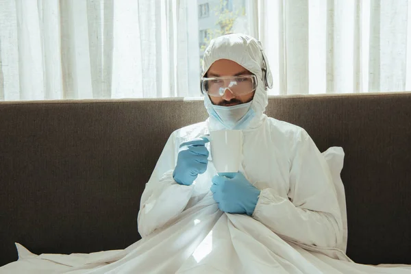Homme en combinaison Hazmat, casque, masque médical, gants en latex et lunettes tenant la tasse au lit — Photo de stock