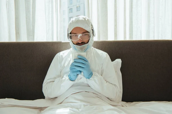 Hombre con traje de materiales peligrosos, auriculares, máscara médica, guantes de látex y gafas que sostienen la taza con bebida en la cama - foto de stock