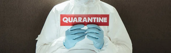 Foto panoramica dell'uomo in attrezzature di protezione individuale in possesso di carta con scritte in quarantena a casa — Foto stock