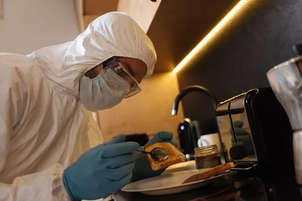 Людина в особистому захисному обладнанні, що тримає ложку біля тостового хліба на кухні — стокове фото