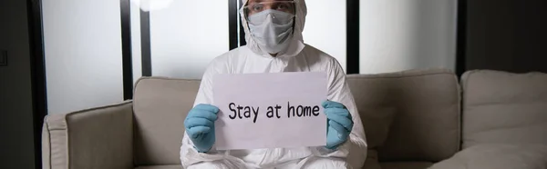 Tiro panorâmico do homem em equipamentos de proteção individual e luvas de látex segurando papel com estadia em casa lettering — Fotografia de Stock