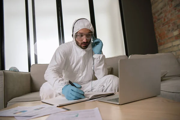 Opérateur barbu en équipement de protection individuelle touchant casque et regardant ordinateur portable dans le salon — Photo de stock