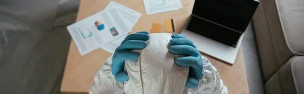 Plano panorámico del hombre en equipo de protección personal tocando la cabeza cerca del ordenador portátil con pantalla en blanco y tarjetas de crédito - foto de stock