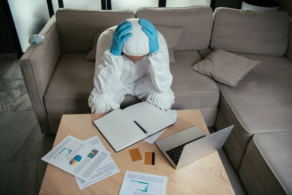 Hombre en equipo de protección personal tocando la cabeza cerca de la computadora portátil, portátil en blanco y tarjetas de crédito - foto de stock