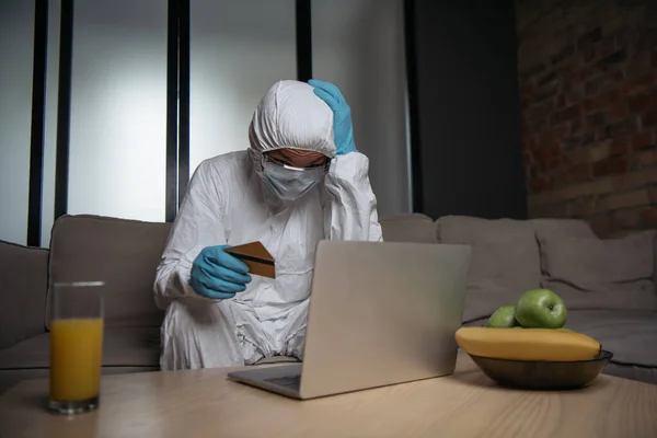 Homem em equipamentos de proteção individual e máscara médica usando laptop e segurando cartões de crédito perto de frutas e vidro de suco de laranja — Fotografia de Stock