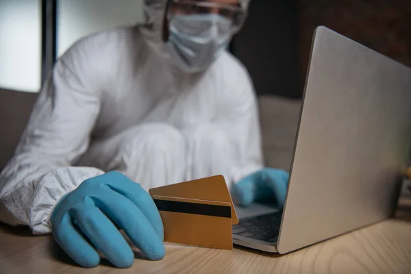 Foyer sélectif de l'homme en équipement de protection individuelle et masque médical à l'aide d'un ordinateur portable et tenant des cartes de crédit — Photo de stock