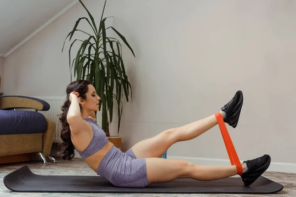 Chica deportiva haciendo abdominales y entrenamiento con banda de resistencia en la alfombra de fitness en casa durante el autoaislamiento - foto de stock