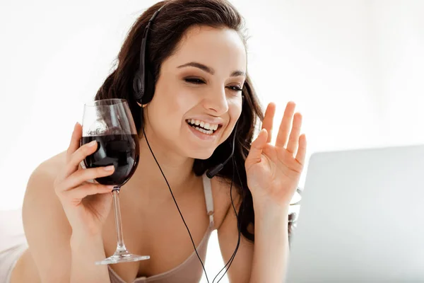 Chica feliz con copa de vino tinto que tiene videollamada con auriculares y portátil durante el autoaislamiento - foto de stock