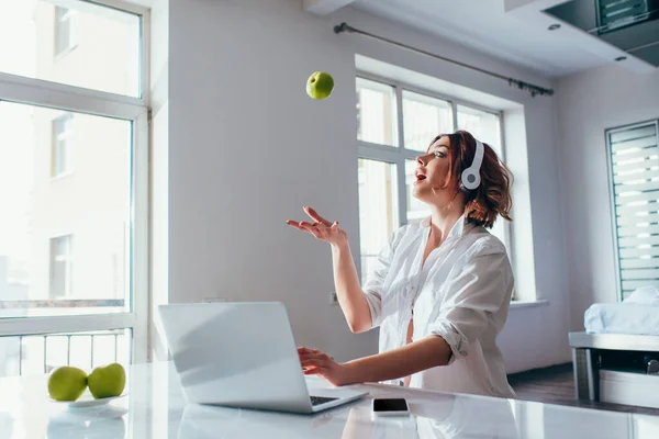 Здивована красива дівчина жонглює з яблуками вдома з цифровими пристроями на карантині — стокове фото