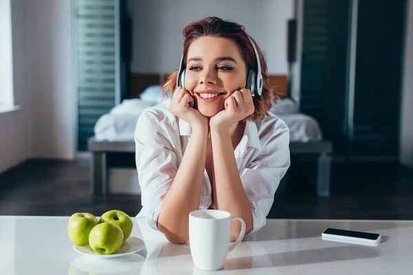 Sorridente ragazza che ascolta musica con cuffie in cucina con caffè, mele e smartphone durante l'autoisolamento — Foto stock