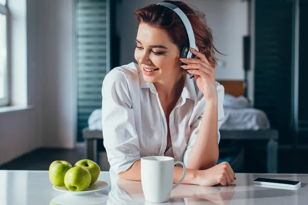 Atractiva chica feliz escuchando música con auriculares en la cocina con café, manzanas y teléfono inteligente durante el autoaislamiento - foto de stock