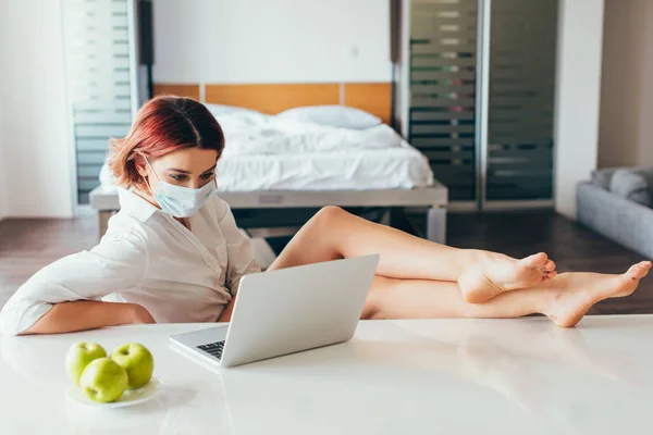 Freelance pieds nus dans le masque médical travaillant sur ordinateur portable à la maison avec des pommes sur l'isolement personnel — Photo de stock