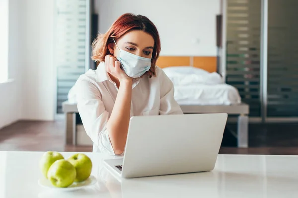 Femme indépendante réfléchie dans un masque médical travaillant sur un ordinateur portable à la maison avec des pommes en quarantaine — Photo de stock