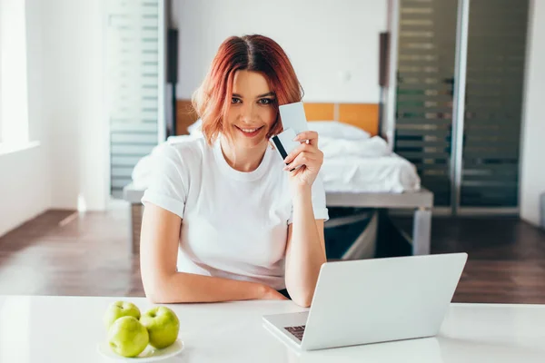 Menina feliz compras on-line com laptop e cartões de crédito na cozinha com maçãs durante a quarentena — Fotografia de Stock
