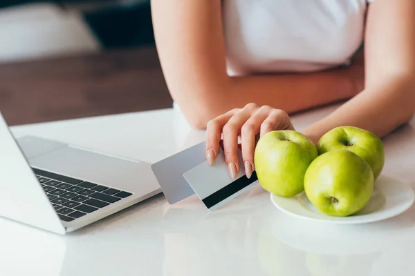 Vue recadrée de femme faisant du shopping en ligne avec ordinateur portable et cartes de crédit sur la cuisine avec des pommes pendant l'isolement personnel — Photo de stock