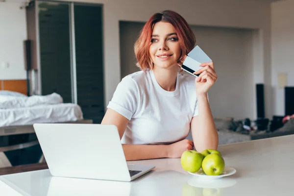 Chica positiva de compras en línea con el ordenador portátil y tarjetas de crédito en la cocina con manzanas durante el autoaislamiento - foto de stock