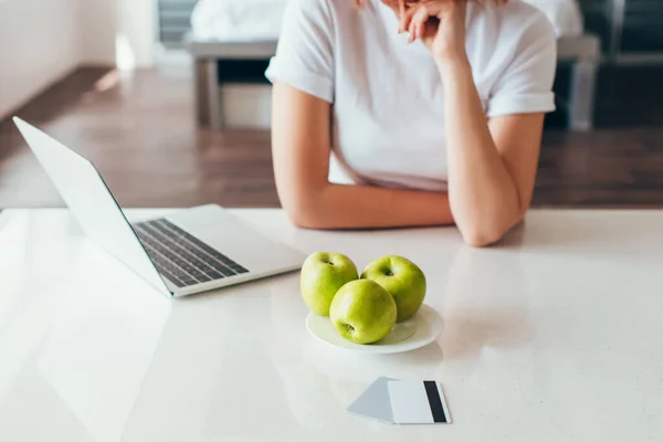 Vista recortada de la chica de compras en línea con el ordenador portátil y tarjetas de crédito en la cocina con manzanas durante la cuarentena - foto de stock