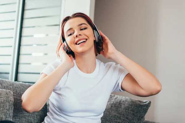 Chica positiva escuchando música con auriculares mientras se sienta en el sofá durante la cuarentena - foto de stock