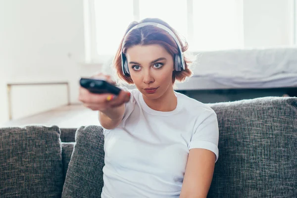 Chica atractiva en auriculares con mando a distancia y viendo la televisión en casa durante el auto aislamiento - foto de stock