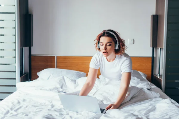 Hermosa chica escuchando música con auriculares y portátil en la cama en el auto aislamiento - foto de stock