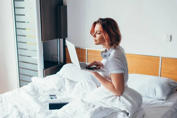 Fille concentrée avec écouteurs à l'aide d'un ordinateur portable au lit avec smartphone et tablette numérique sur l'isolement personnel — Photo de stock