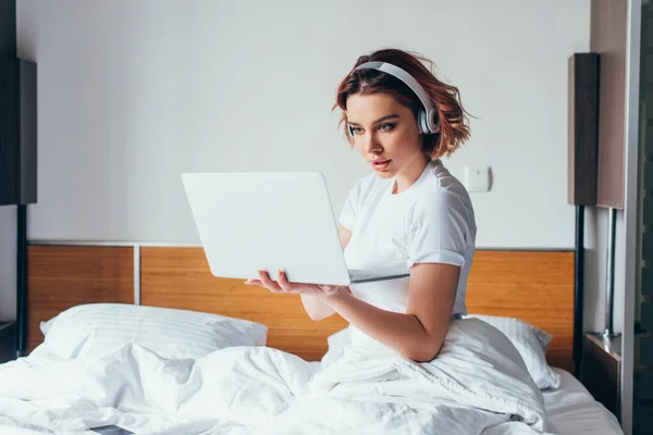 Bella ragazza concentrata in cuffie utilizzando il computer portatile a letto in quarantena — Foto stock