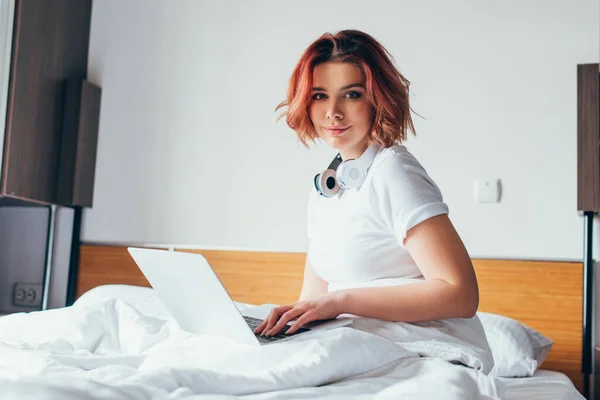 Attraente ragazza con le cuffie utilizzando il computer portatile a letto su auto isolamento — Foto stock