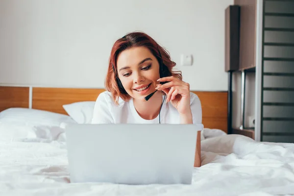 Schöne glückliche Mädchen macht Videoanruf mit Headset und Laptop im Bett während der Selbstisolation — Stockfoto