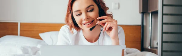 Attraktives Mädchen macht Videoanruf mit Headset und Laptop im Bett während der Selbstisolierung, panoramische Ernte — Stockfoto