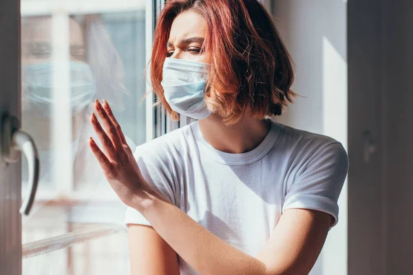 Upset girl in medical mask looking through window during self isolation — Stock Photo