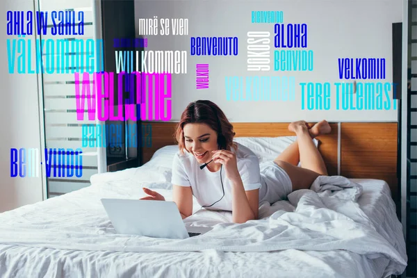 Feliz freelancer haciendo videollamada con auriculares y portátil en la cama durante el autoaislamiento con letras - foto de stock