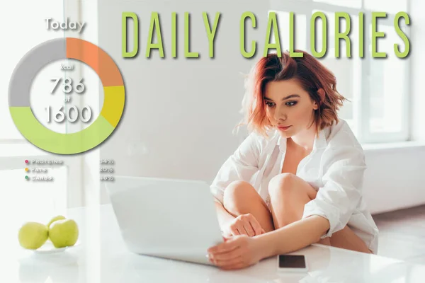 Attractive girl with headphones using laptop on kitchen with apples and smartphone during self isolation with daily calories lettering — Stock Photo