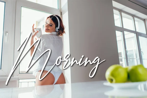 Happy girl listening music in headphones and holding coffee cup on kitchen with apples during self isolation with morning lettering — Stock Photo