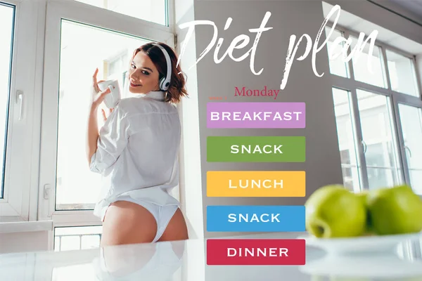 Happy girl listening music in headphones and holding coffee cup on kitchen with apples during self isolation with diet plan lettering — Stock Photo