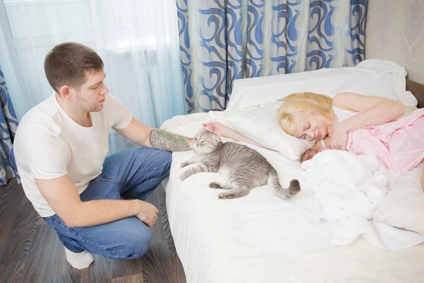 Portret van mooie jonge ouders en schattige baby liggen samen in bed. Een schattige grijze kat zit naast hen. Stockafbeelding