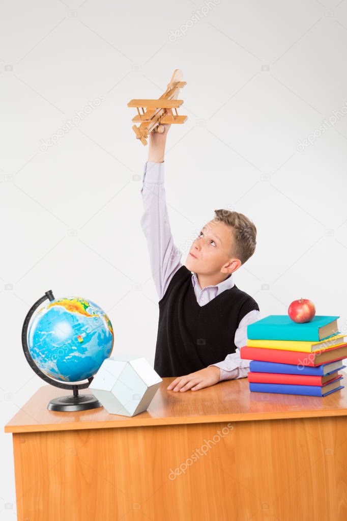 Cute schoolboy plays by airplane at a school desk isolated on white background