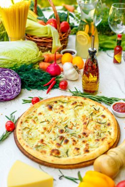 Tahta bir yüzeyde soğanlı ve fesleğenli sıcak pizza ve beyaz bir masada sebzeler.