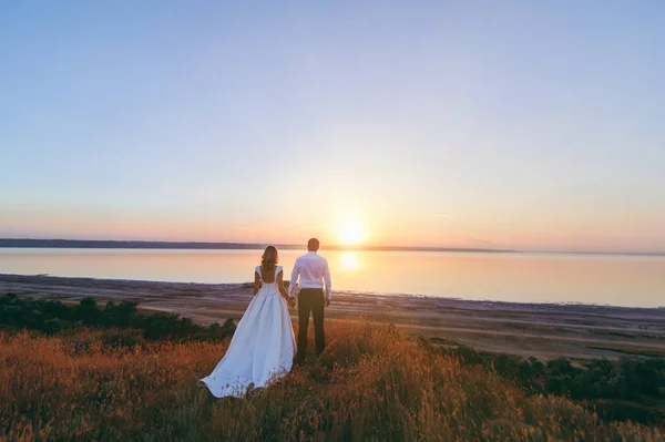 Mariée et marié marchent ensemble au coucher du soleil près de la mer — Photo