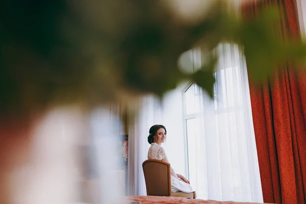 Sposa bruna in velo e raso bianco vestaglia siede vicino alla finestra — Foto Stock