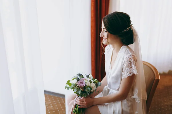 Bride brunette dengan kerudung dan gaun satin putih duduk di dekat jendela — Stok Foto