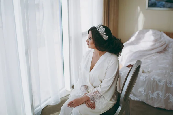 Νύφη μελαχρινή σε λευκό σατέν ρόμπα κάθεται από το παράθυρο — Φωτογραφία Αρχείου